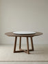 Isolated view of a round walnut dining table with trestle base and centered white marble lazy Susan on a woven rug.