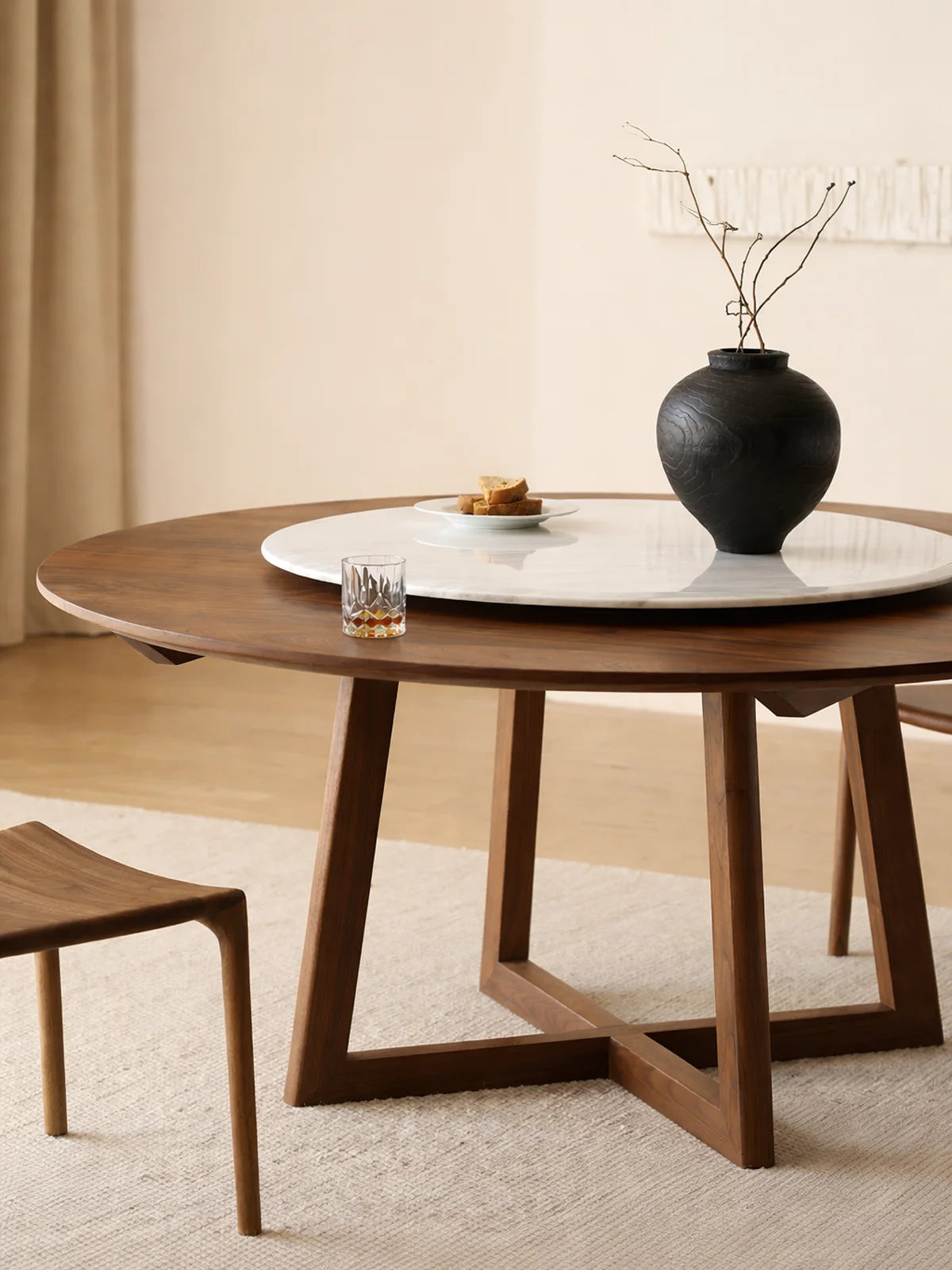 Three-quarter view of a round walnut dining table with angled trestle base and large white marble lazy Susan.