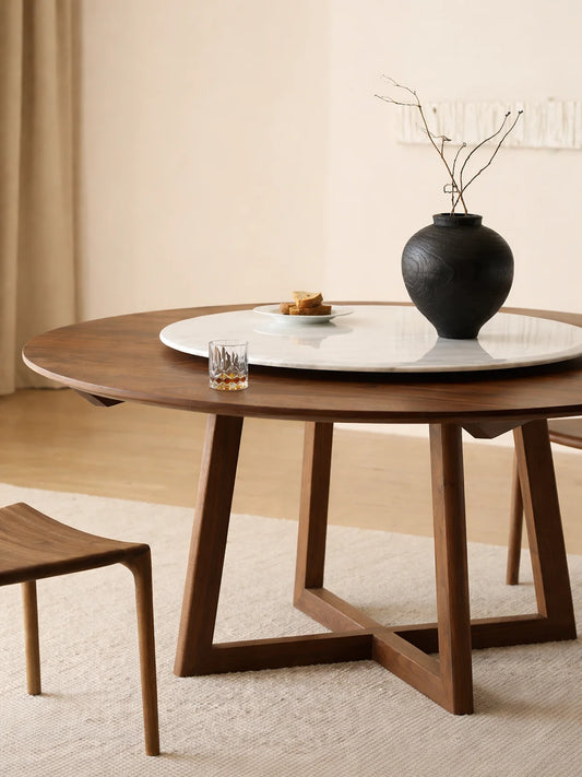 Three-quarter view of a round walnut dining table with angled trestle base and large white marble lazy Susan.