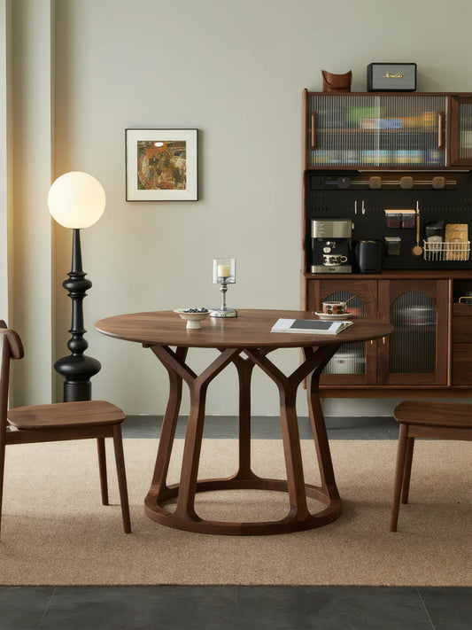 Front view of a round solid walnut dining table with an open sculptural base, styled in a warm modern interior.