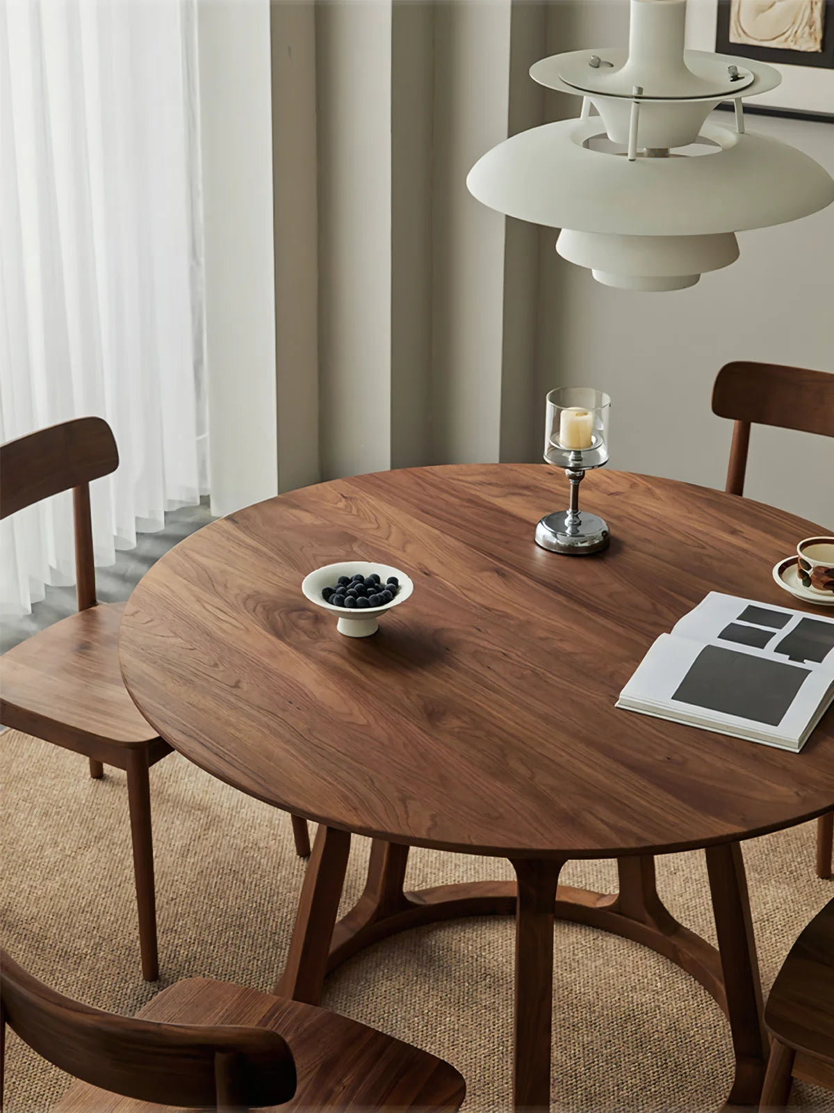 Close view of a round solid walnut dining table showing the softly eased top, rich wood grain, and open pedestal base in a calm dining setting.