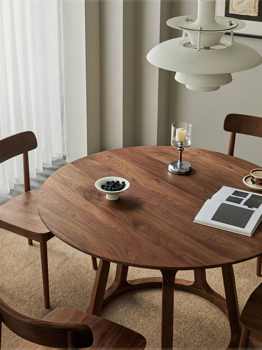 Close view of a round solid walnut dining table showing the softly eased top, rich wood grain, and open pedestal base in a calm dining setting.