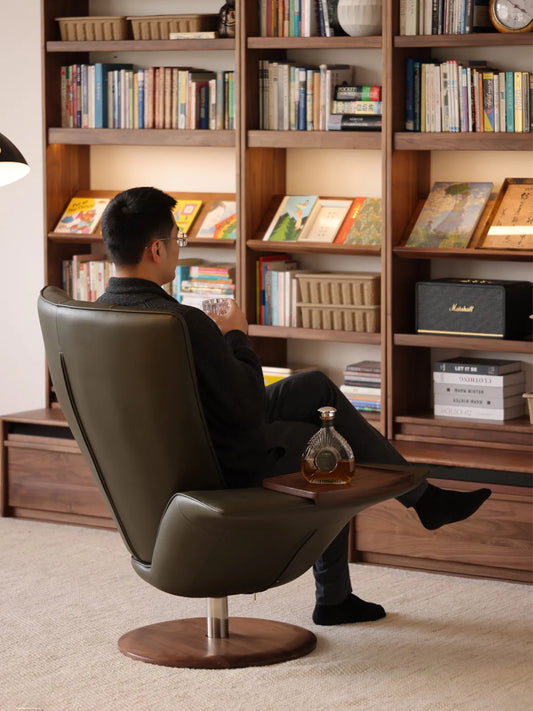 A man relaxing in The Helios Retreat Swivel Recliner in a home library, utilizing the attached black walnut side tray for his drink.