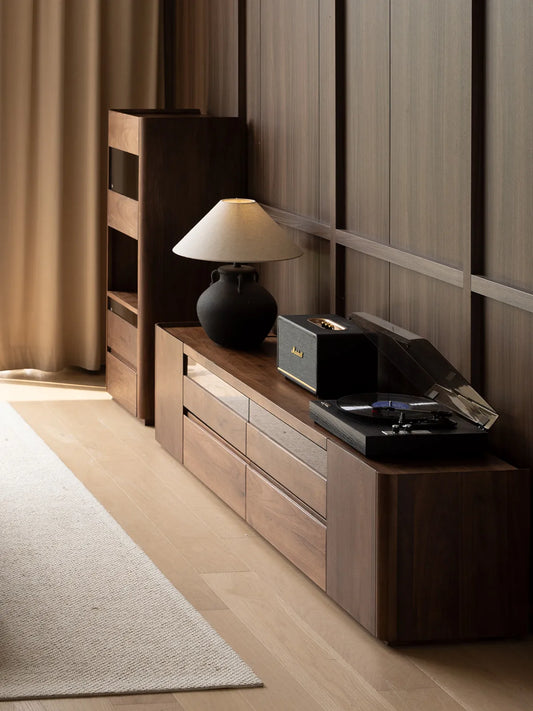 Angled view of The Pebble Media Console in natural walnut, styled with a record player and lamp against a dark wood-paneled wall.