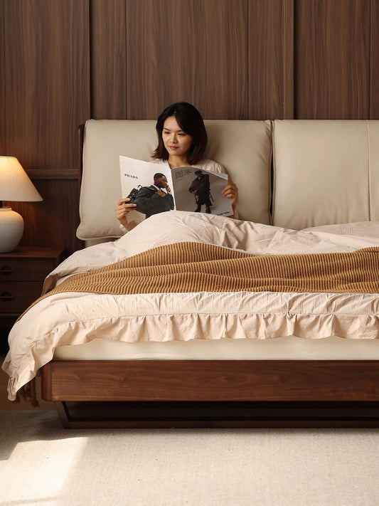 A woman relaxing and reading comfortably in the spacious walnut king bed frame, leaning against the soft and supportive headboard.