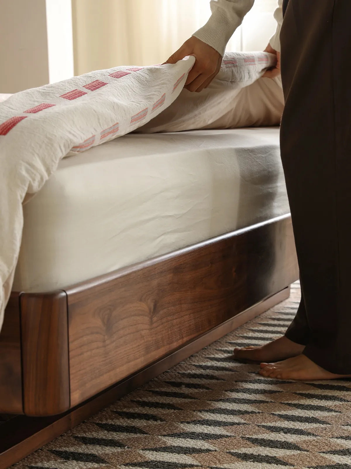 A person tucking in the sheets on the walnut queen bed frame, showcasing the smooth solid wood side rails and secure mattress inset.