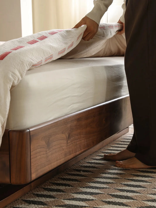 A person tucking in the sheets on the walnut queen bed frame, showcasing the smooth solid wood side rails and secure mattress inset.