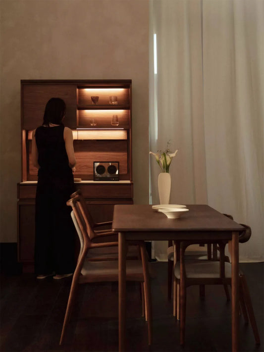 Detail of the Walnutry walnut wood dining table showing the rich matte wood grain finish and warm styling, with a woman standing beside it for scale.