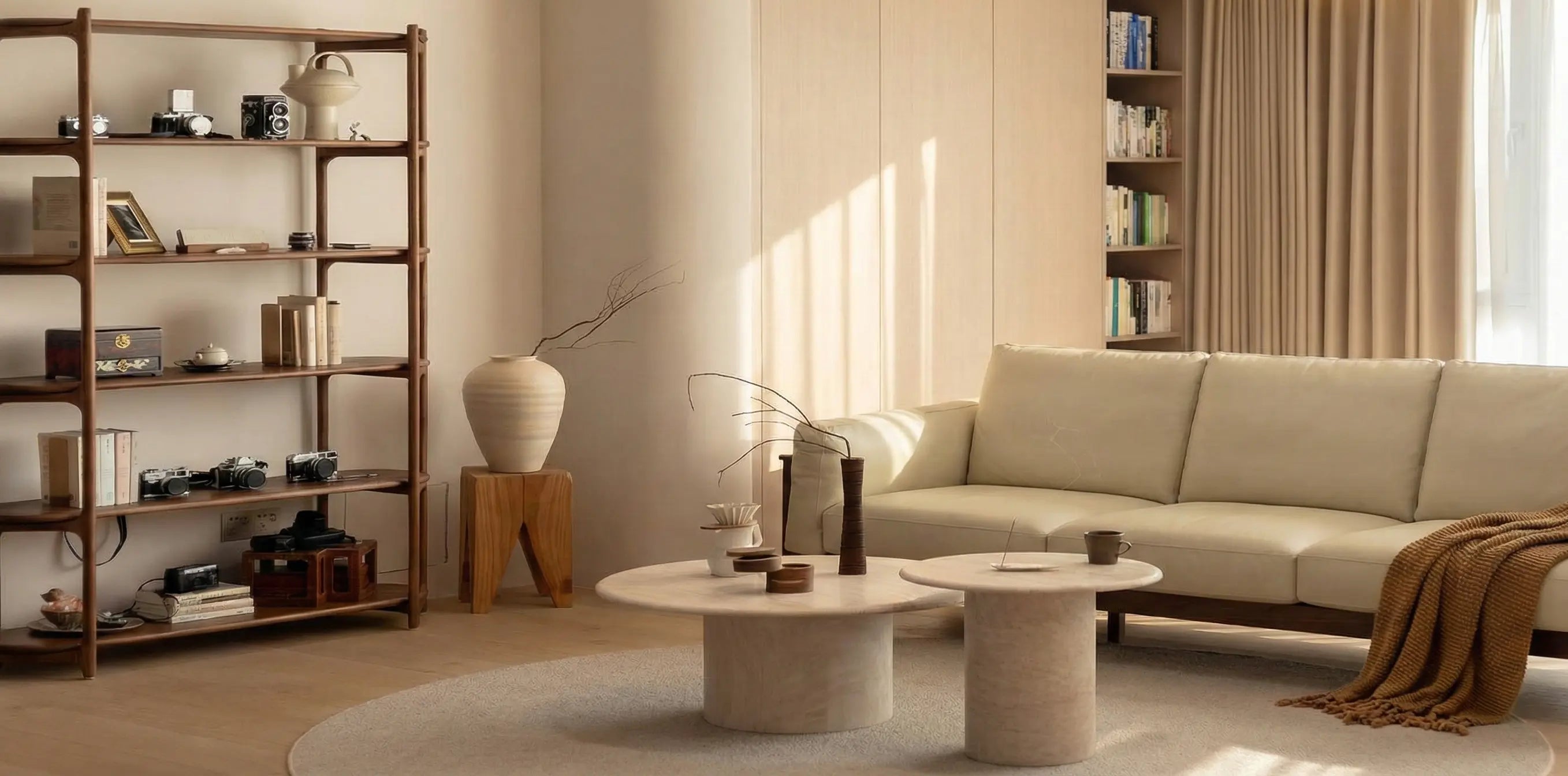 A sun-drenched living room featuring a tall solid walnut open shelving unit filled with books and vintage cameras. A cream sofa sits to the right with round travertine coffee tables in the center, creating a calm, natural atmosphere.